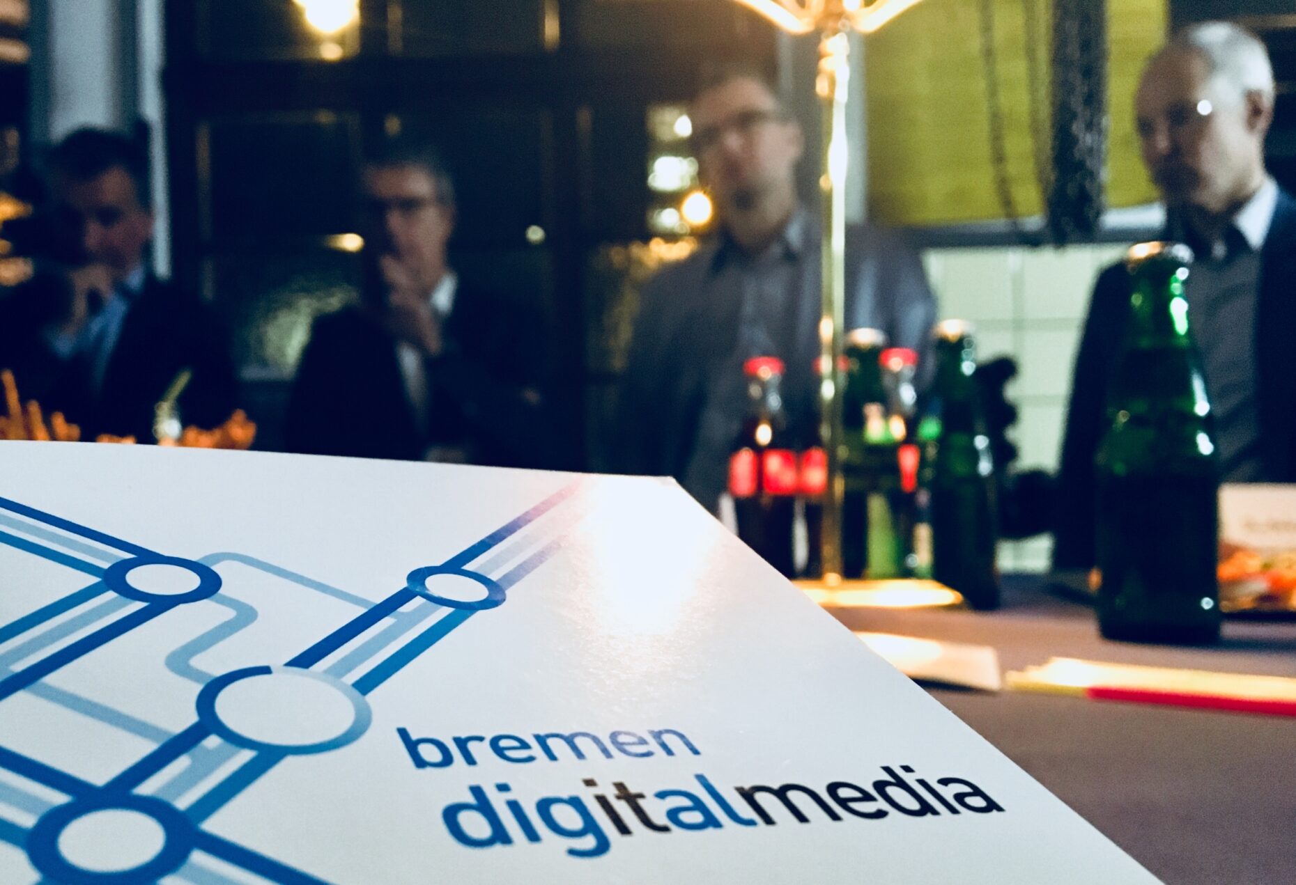 Ein Flyer von bremen digitalmedia ist im Bild-Vordergrund; dahinter sieht man verschwommen Menschen, die um einen Tisch stehen.