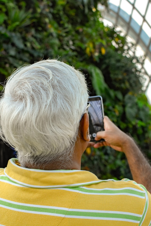 Eine Person mit Grauen Haaren ist von hinten zu sehen und hält ein Smartphone in der Hand.