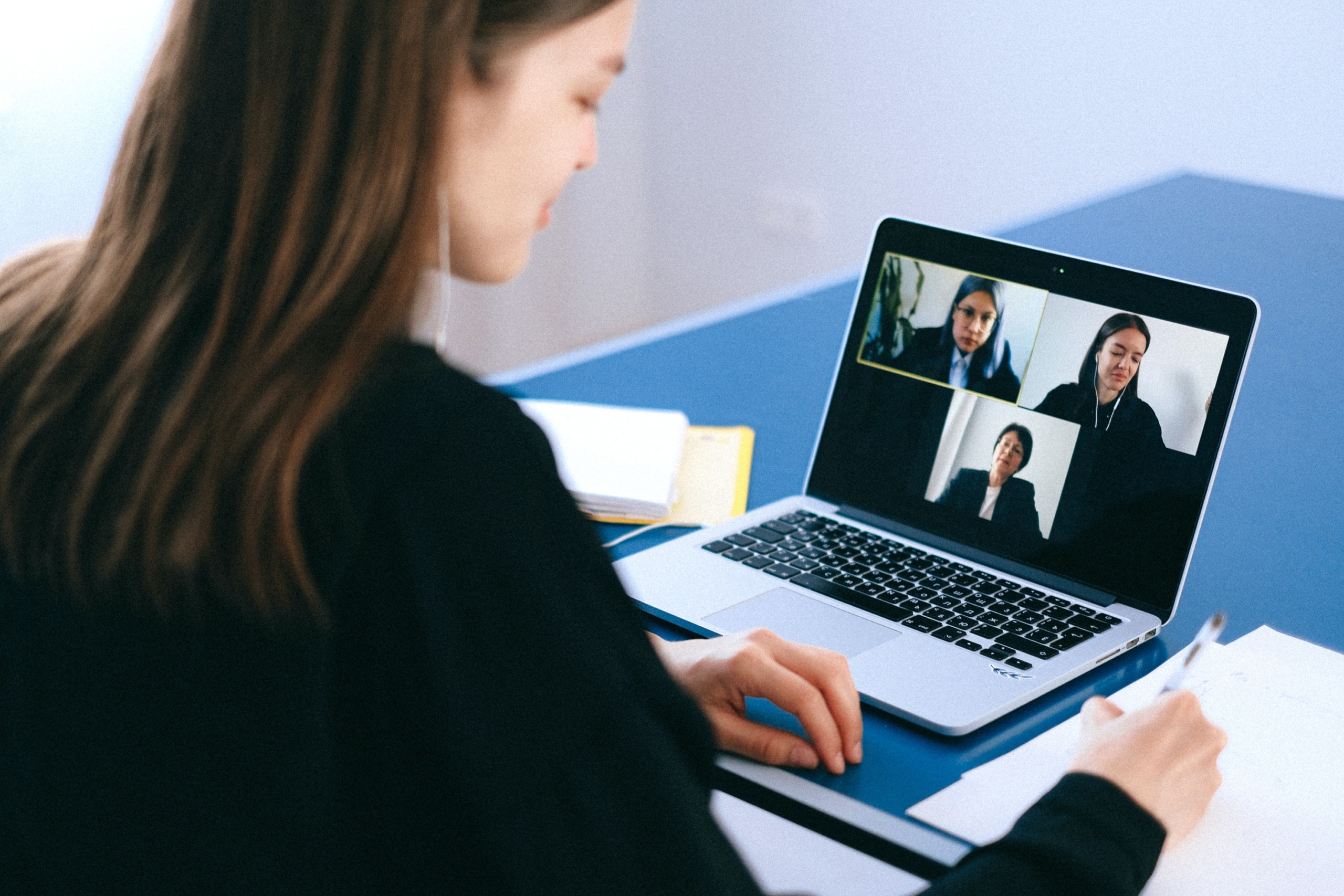 Eine Frau sitzt seitlich mit dem Rücken zur Kamera und schaut auf einen Laptop, der drei Personen zeigt, die gerade in einem Meeting mit ihr sind.