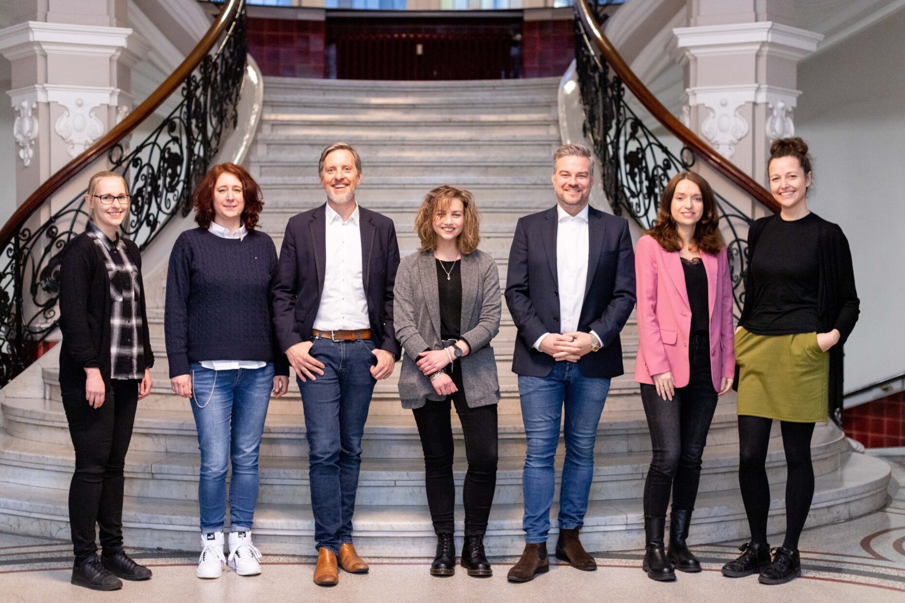 Sieben Menschen stehen im Foyer eines alten, gut restaurierten Hauses vor einer großen Treppe und lächeln in die Kamera.