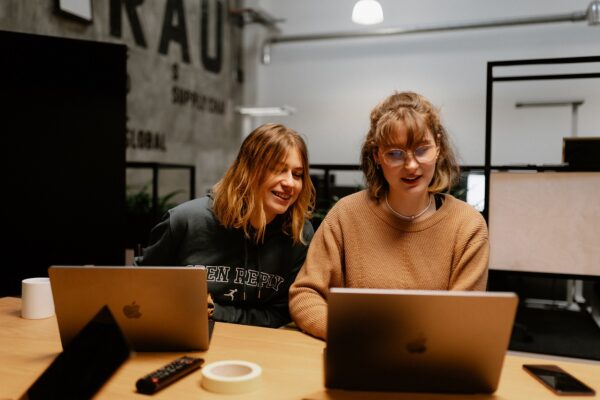 Zwei Frauen sitzen hinter einem großen Holztisch und schauen gemeinsam auf einen aufgeklappten Laptop.