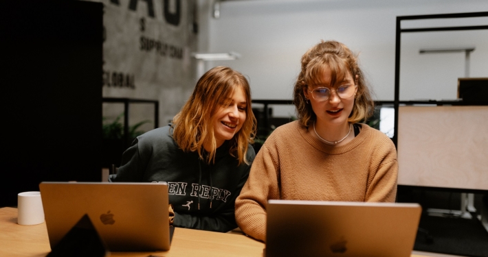 Zwei Frauen sitzen hinter einem großen Holztisch und schauen gemeinsam auf einen aufgeklappten Laptop.