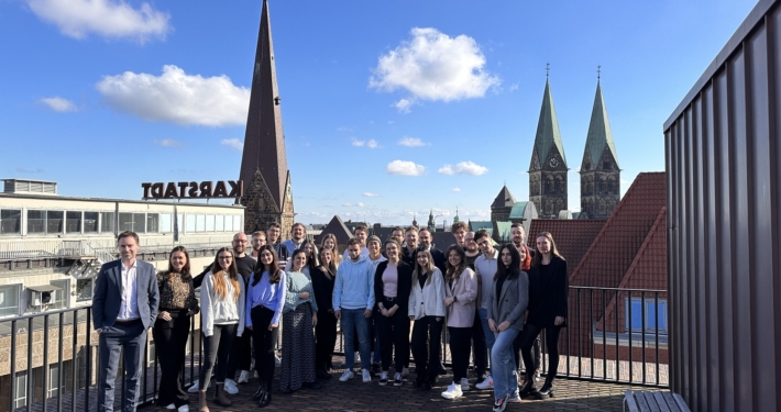 Eine Gruppe von Menschen steht auf der Dachterasse der Stadt Bremen und posiert für ein Gruppenfoto. Im Hintergrund kann man Dächer und Kirchtürme erkennen.