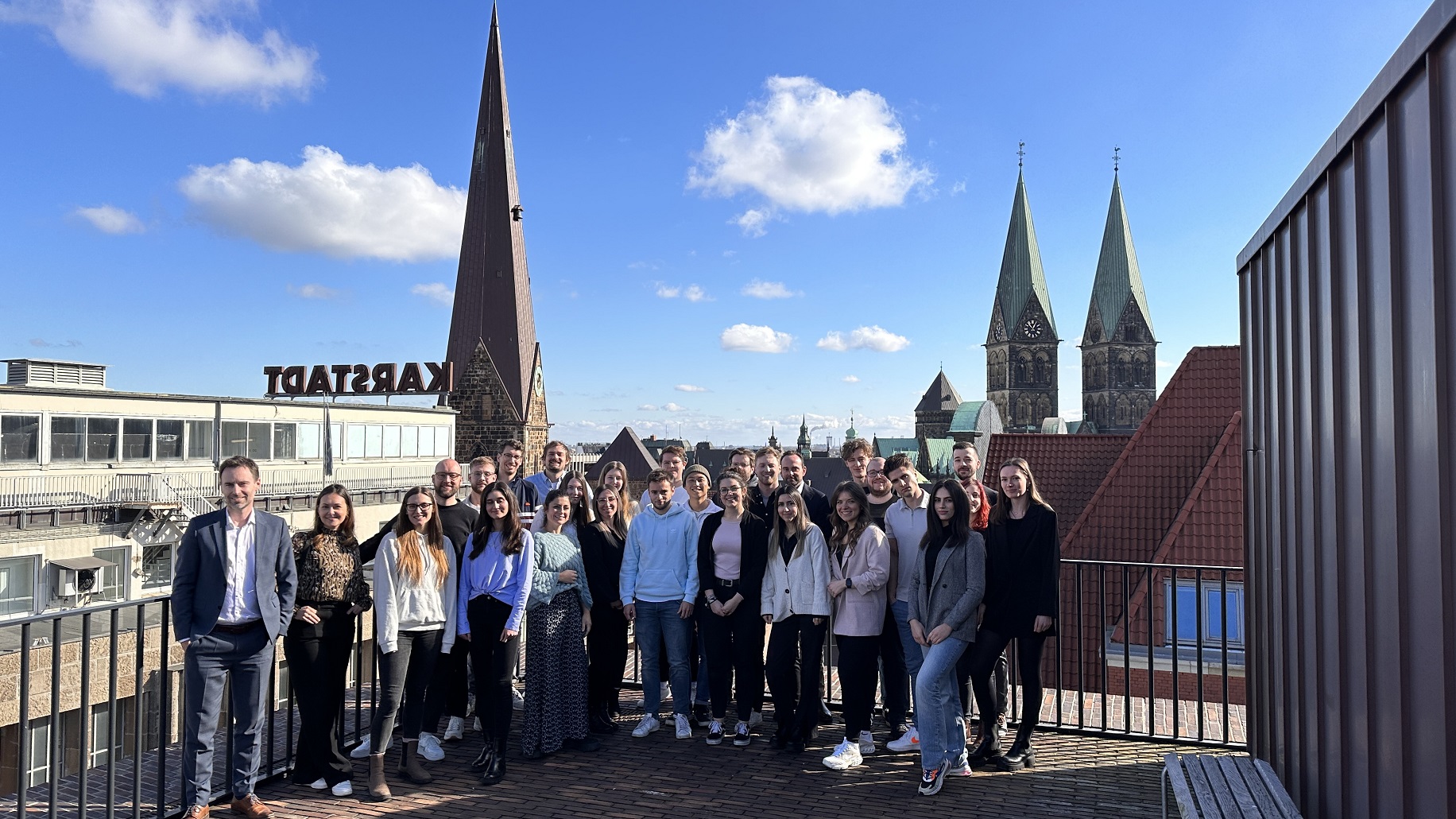 Eine Gruppe von Menschen steht auf der Dachterasse der Stadt Bremen und posiert für ein Gruppenfoto. Im Hintergrund kann man Dächer und Kirchtürme erkennen.