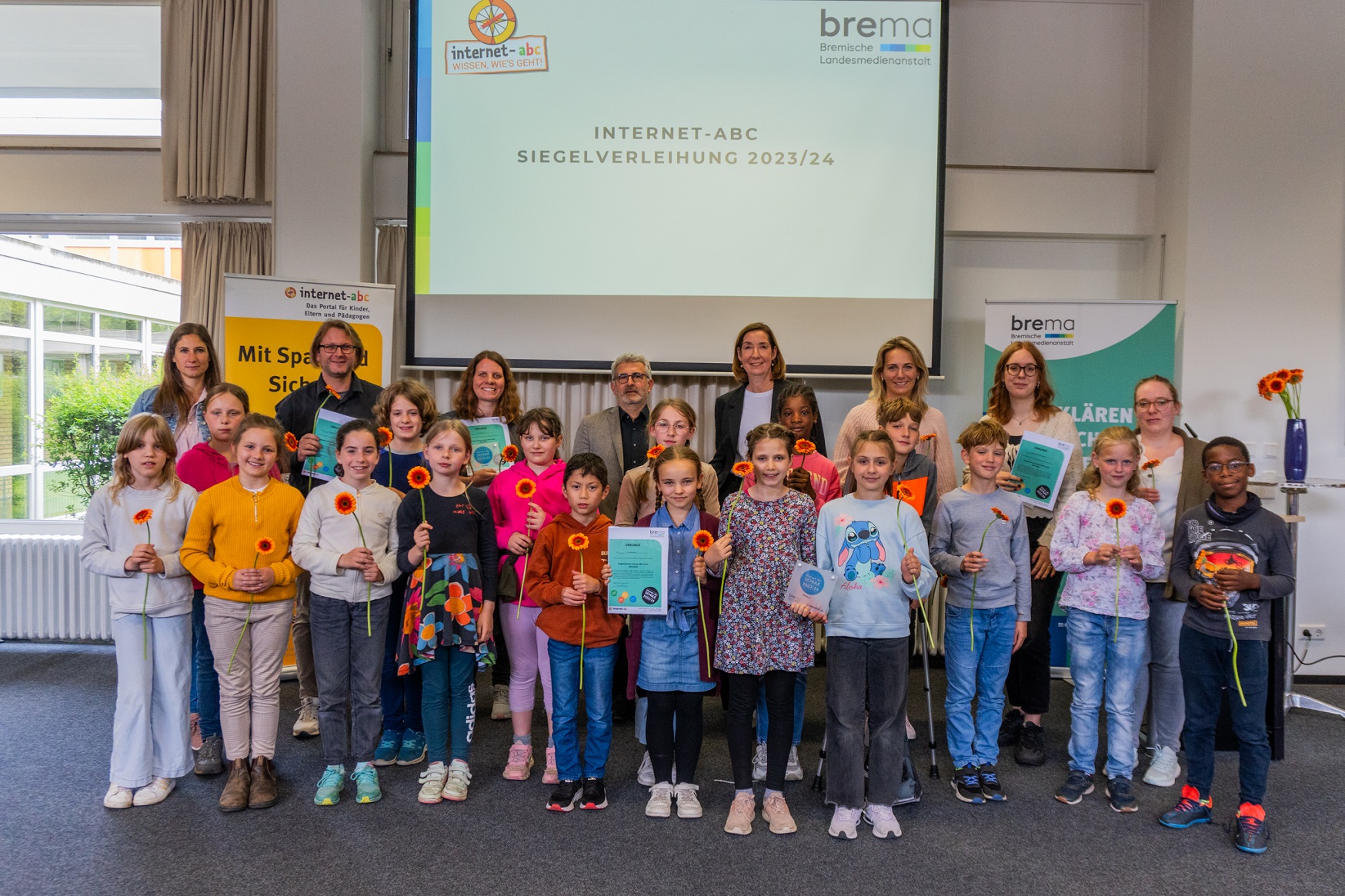 Eine Gruppe von Schülerinnen und Schülern sowie einigen Erwachsenen halten einen Urkunde und einen orangefarbene Blume in der Hand und posieren für ein Gruppenfoto.