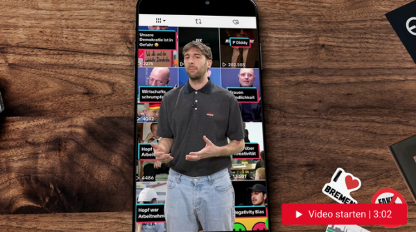 Dunkelhaariger Mann steht vor einem großen Handy, auf dem der TikTok-Feed zu sehen ist.