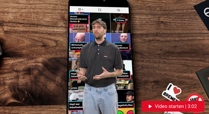 Dunkelhaariger Mann steht vor einem großen Handy, auf dem der TikTok-Feed zu sehen ist.