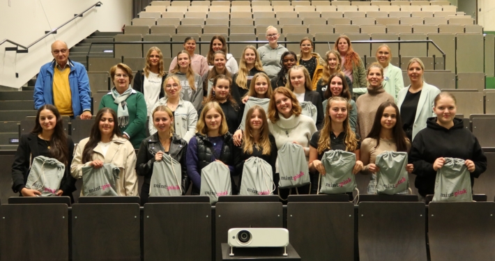 27 Schülerinnen stehen in einem Hörsaal. Sie werden begleitet von einem Mann (links stehend) und einer erwachsenen Frau (rechts stehend).