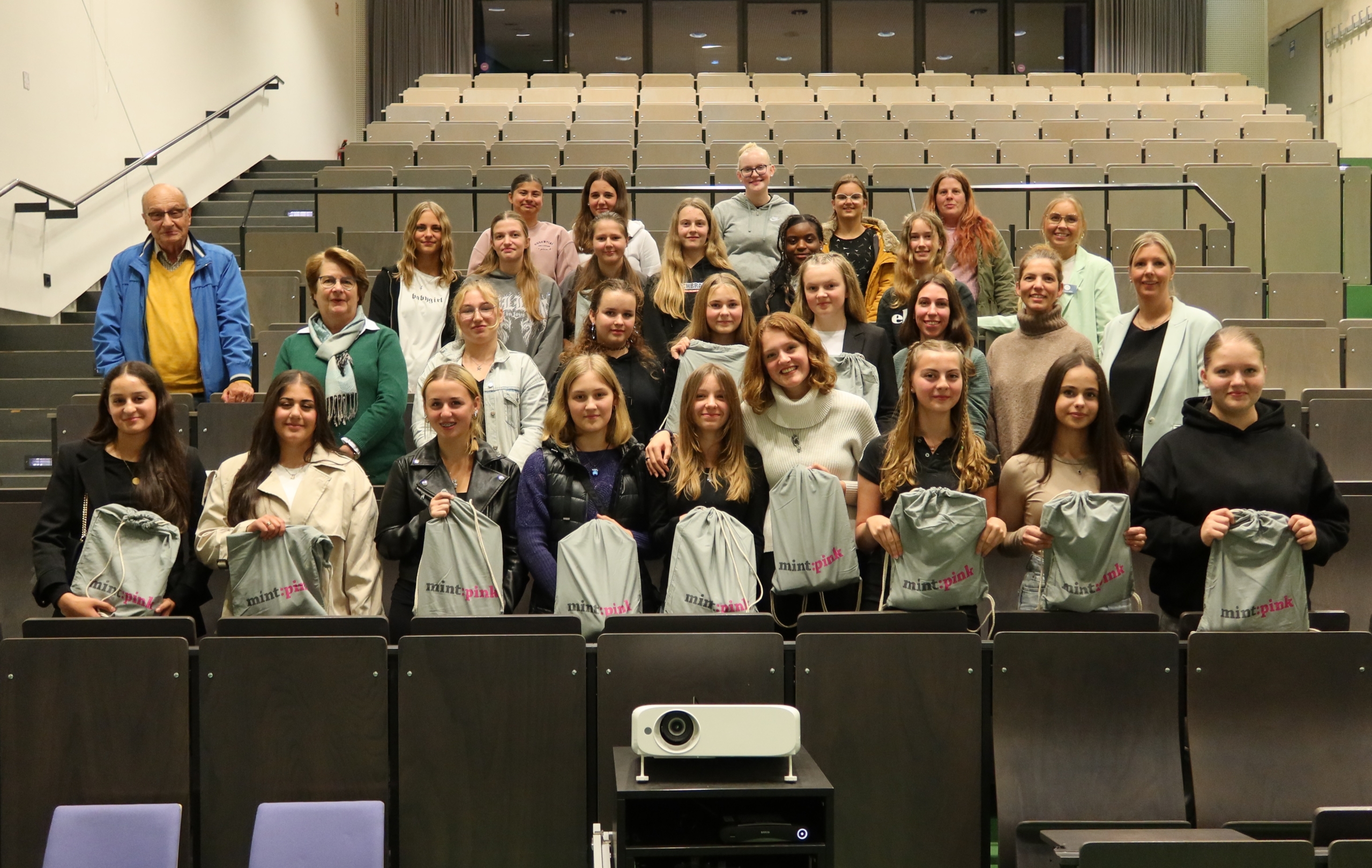 27 Schülerinnen stehen in einem Hörsaal. Sie werden begleitet von einem Mann (links stehend) und einer erwachsenen Frau (rechts stehend).