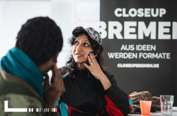 Eine Frau sitzt am Tisch und schaut nach links oben aus dem Foto. Ihr gegenüber und mit dem Rücken zur Kamera sieht man eine weitere Person sitzen.