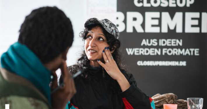 Eine FRau sitzt am Tisch und schaut nach links oben aus dem Foto. Ihr gegenüber und mit dem Rücken zur Kamera sieht man eine weitere Person sitzen.