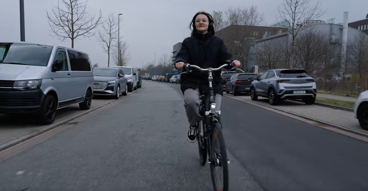 Ein Screenshot eines Videos. Eine Frau fährt auf einem Fahrrad durch eine Straße.