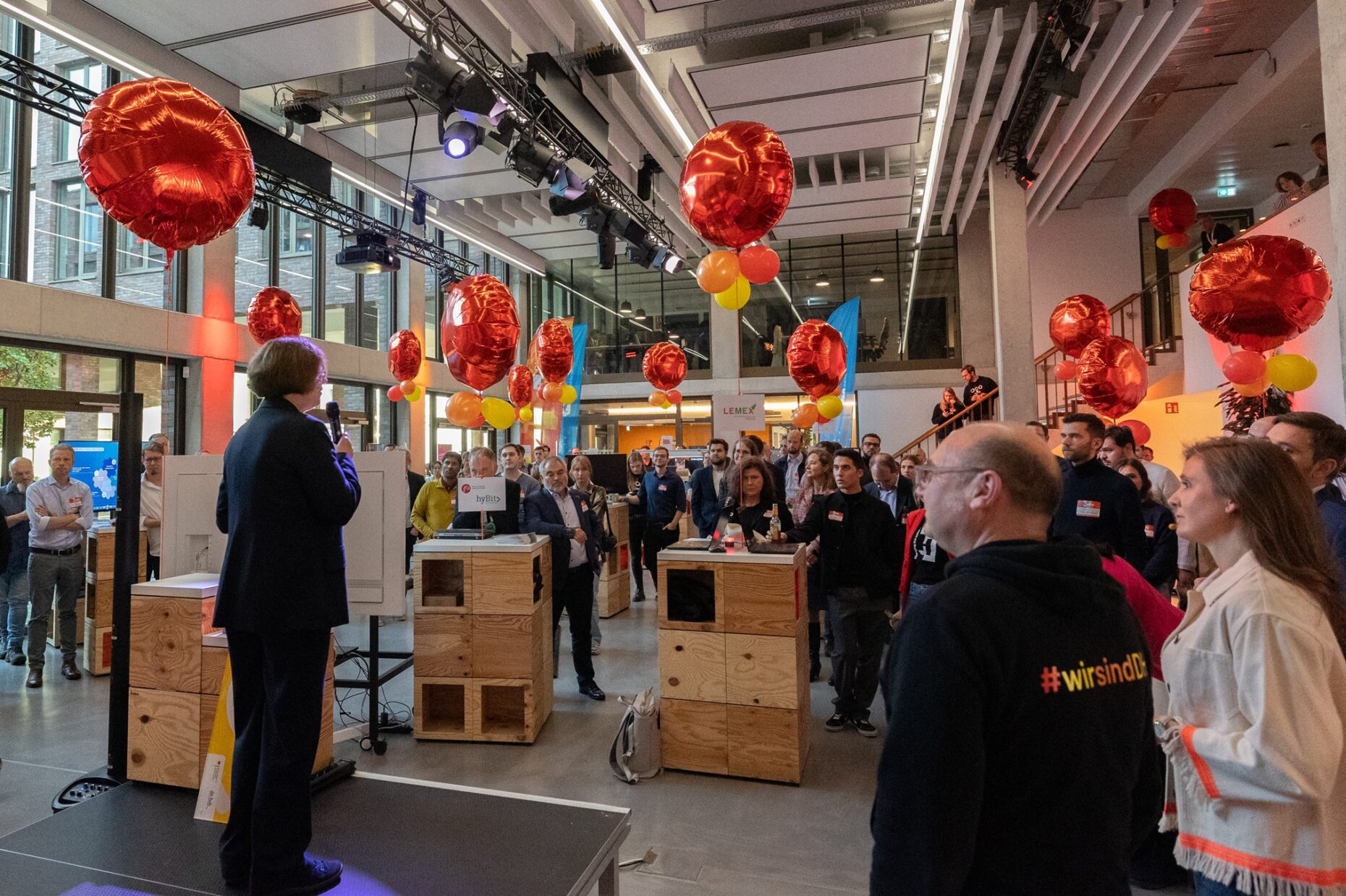 Viele Personen stehen um eine kleine Bühne herum, auf der eine Person mit Mikro steht. In der Luft hängen Luftballons.