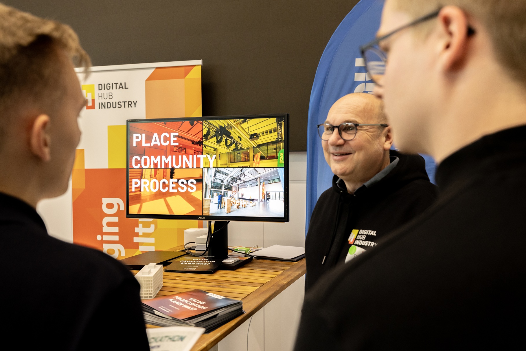 Drei Personen stehen an einem Messestand mit Rollups und einem großen Bildschirm.