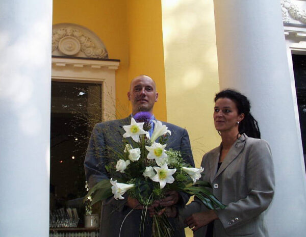 Zwei Personen stehen mit einem großen Blumenstrauß vor einem Gebäude.